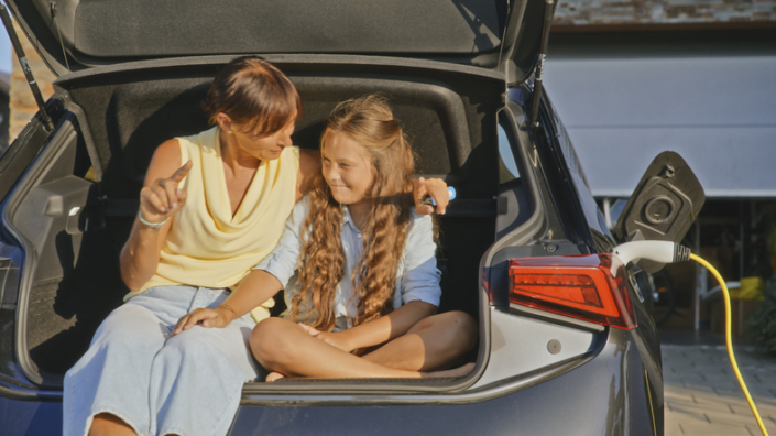 Mother and Daughter Relaxing in Electric Car Trunk While Charging in Front of Modern Home